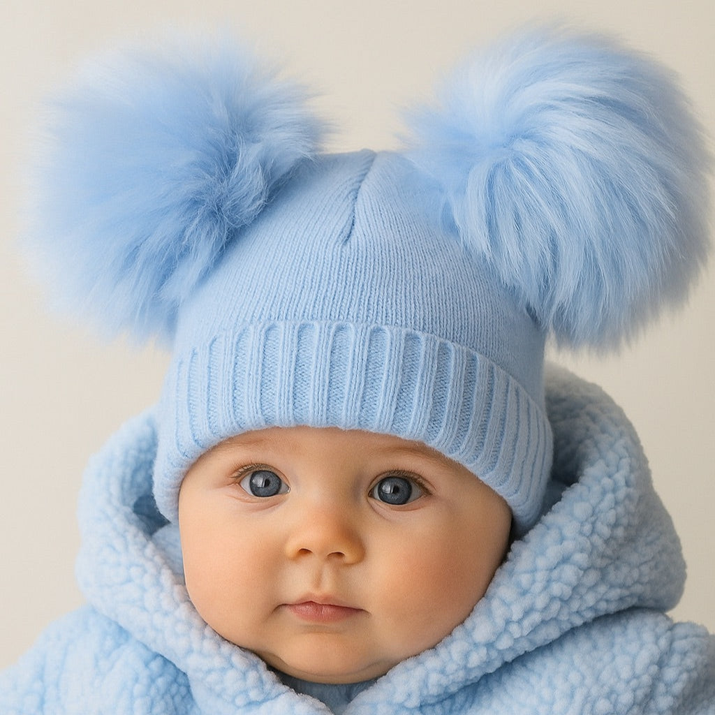 Baby wearing a light blue knit hat with pom-poms and matching coat against a neutral background
